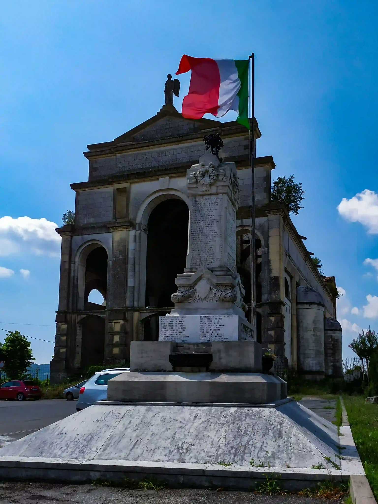 Chiesa incompiuta sullo sfondo, in primo piano monumento ai caduti con bandiera italiana
